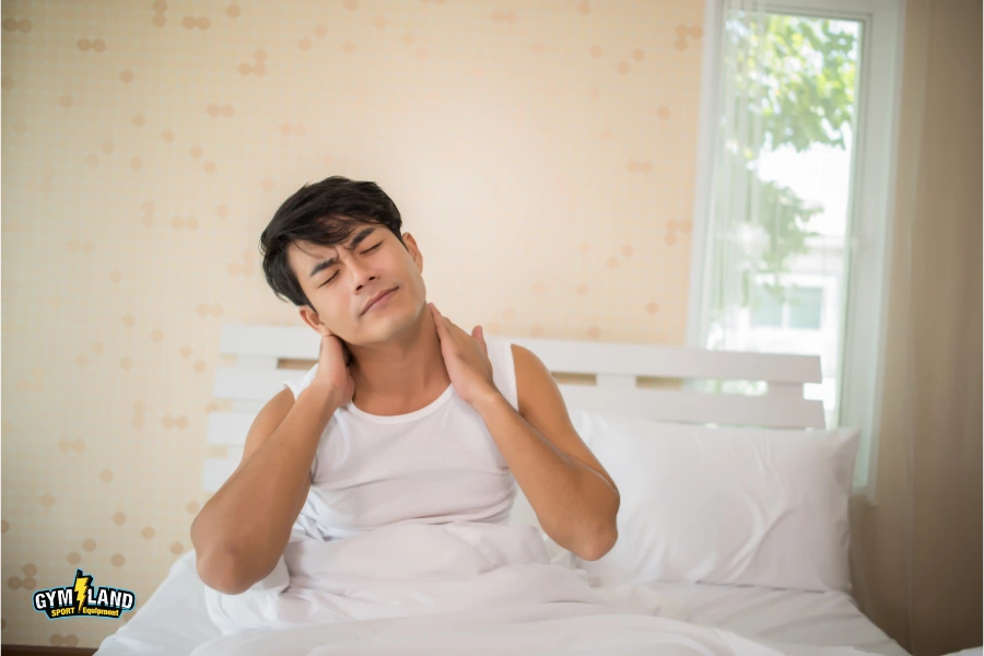 مردی آسیایی با زیرپیراهنی سفید روی تخت خواب سفید که با دو دست، گردنش را گرفته و حالت خواب آلوده دارد.